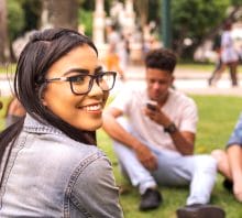Young brazilian woman enjoying time with her friends at the park. Multi racial group of friends sitting on the grass. Focus on the rear viewed girl smiling at camera. There are people on the blurred background.